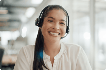A smiling woman wearing a headset with a microphone looks engaged while working in a bright office setting.
