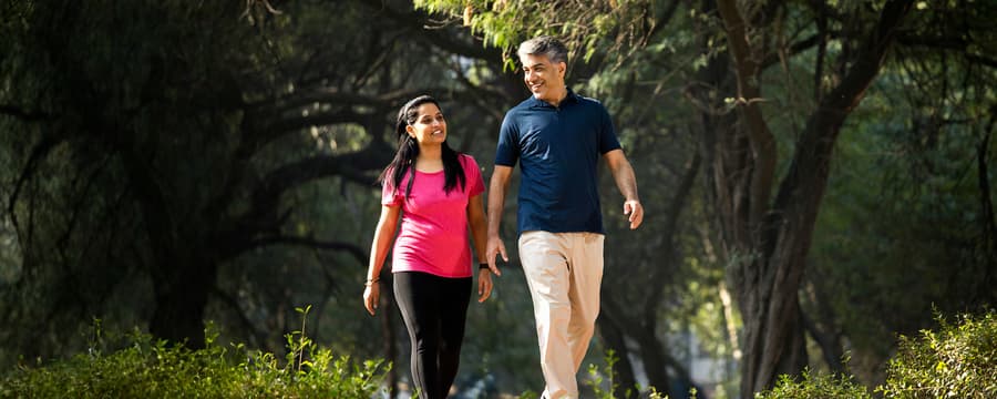 A smiling man and woman walking together on a sunlit path surrounded by trees and greenery.