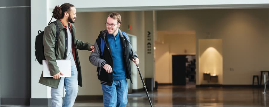Two people walk together through a spacious indoor building, smiling as they talk. One person carries a white laptop and backpack, while the other uses a white cane and wears a backpack, highlighting an accessible and supportive environment.