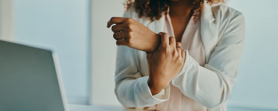 Woman seated at a desk holds her wrist while working on a laptop, suggesting wrist or hand discomfort.