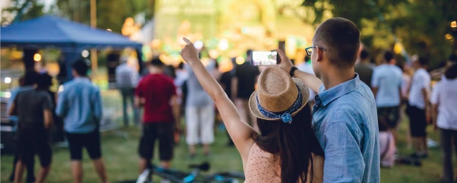 A couple stands arm in arm at an outdoor concert, taking a selfie with a brightly lit stage in the background while a crowd gathers on the grass around them.