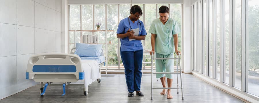 Healthcare professional walks beside and assists a barefoot patient using a walker in a bright hospital rehabilitation hallway.