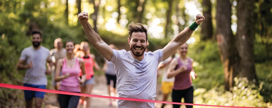 Smiling runner raises his arms in celebration as he crosses the finish line during a race on a wooded trail.