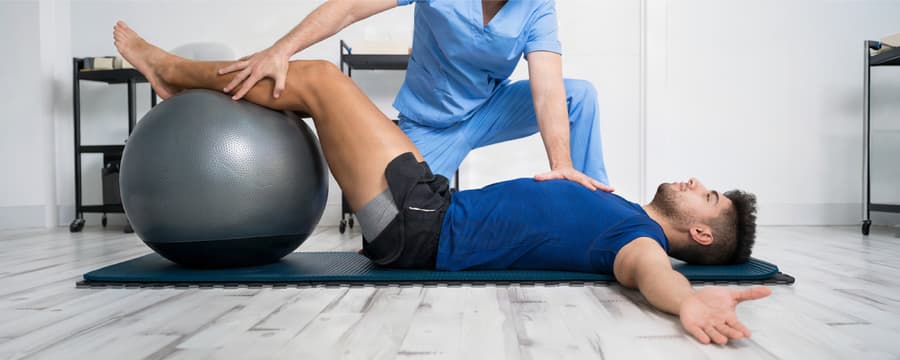 Physical therapist assists a man lying on an exercise mat with his legs elevated on a stability ball during a pelvic floor strengthening exercise.