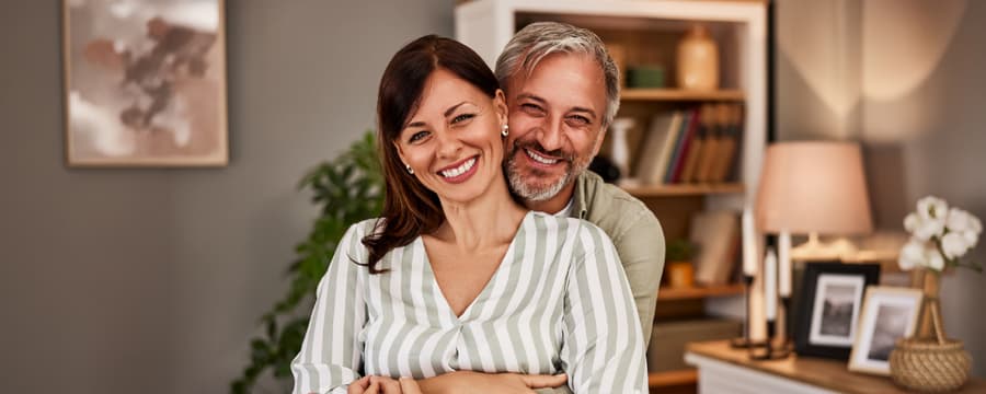 Smiling middle-aged couple embracing in a cozy, modern living room, with the man hugging the woman from behind as both look at the camera. A bookshelf, houseplant, and warm home décor create a welcoming background.