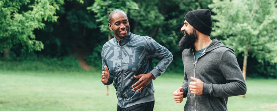Two adults jogging side by side on a paved path in a green park, smiling and talking as they run. One wears a patterned gray athletic jacket and black leggings, while the other wears a dark beanie, gray hoodie, and blue shorts. Trees and grass fill the background.