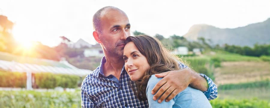 Middle-aged couple embracing outdoors, with the woman resting her head on the man's shoulder as he holds her close.