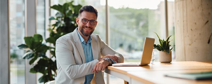 A smiling man wearing glasses and a light-colored blazer sits at a desk with an open laptop in a bright, plant-filled office.