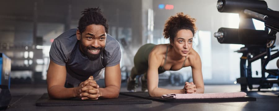 A man and woman hold plank positions on exercise mats while working out together in a gym.