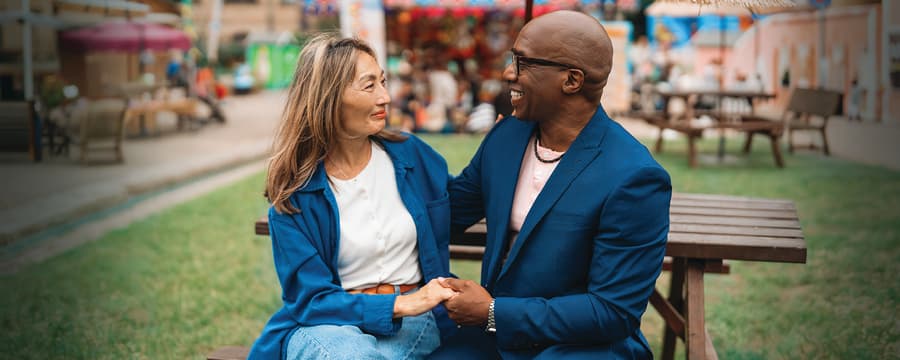 Two people sitting closely together outdoors, smiling and holding hands while talking. One person wears a blue blazer and glasses, and the other wears a blue jacket over a white shirt. A blurred park or market scene appears in the background.