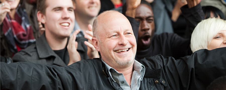 A bald man in a black jacket and light blue shirt stands at the center of a smiling group of diverse people, his arms raised in celebration. The crowd behind him appears joyful and supportive, suggesting a moment of community encouragement or shared accomplishment.