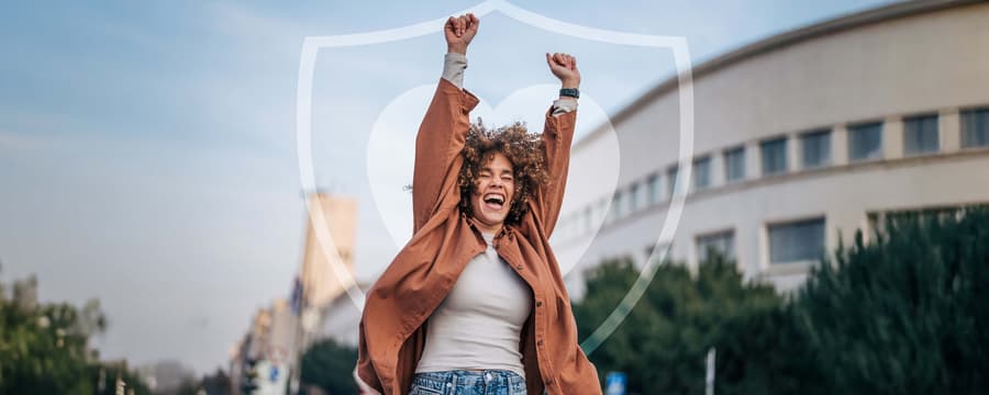 Smiling person outdoors with arms raised in celebration in front of a subtle heart-and-shield graphic, symbolizing confidence and protection.
