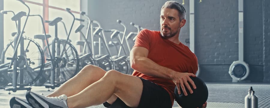 A man in a red athletic shirt and black shorts performs a seated core exercise on a gym mat, twisting his torso while holding a medicine ball. Exercise equipment like stationary bikes is visible in the background, and a towel lies on the floor nearby.