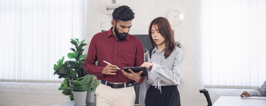 Two coworkers stand in a bright office, reviewing information together on a tablet while one holds a stylus.