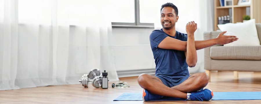 Smiling man sitting cross-legged on an exercise mat and stretching his arm during a home workout.
