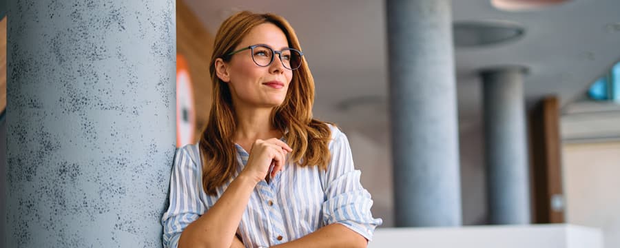 Woman wearing glasses leans against a column in a modern office, looking thoughtfully into the distance.