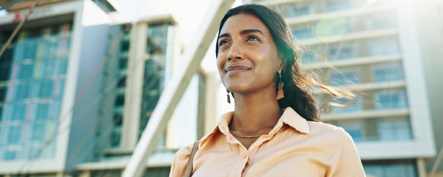 A woman in a light blouse stands outdoors in front of modern buildings, smiling slightly and looking ahead, with sunlight creating a warm glow around her.
