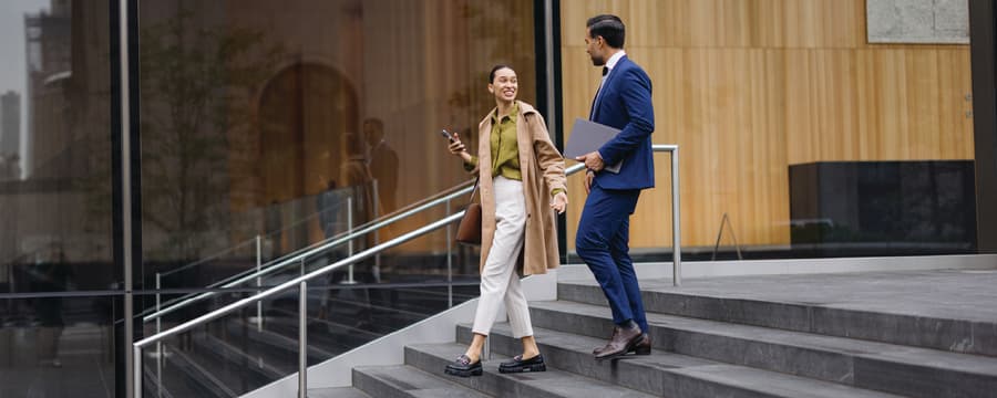 Two young professionals talking while walking down outdoor steps of a modern office building, one holding a smartphone and the other carrying a laptop.