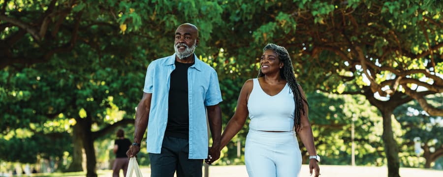 Smiling couple holding hands while walking together on a tree-lined path in a sunny park.
