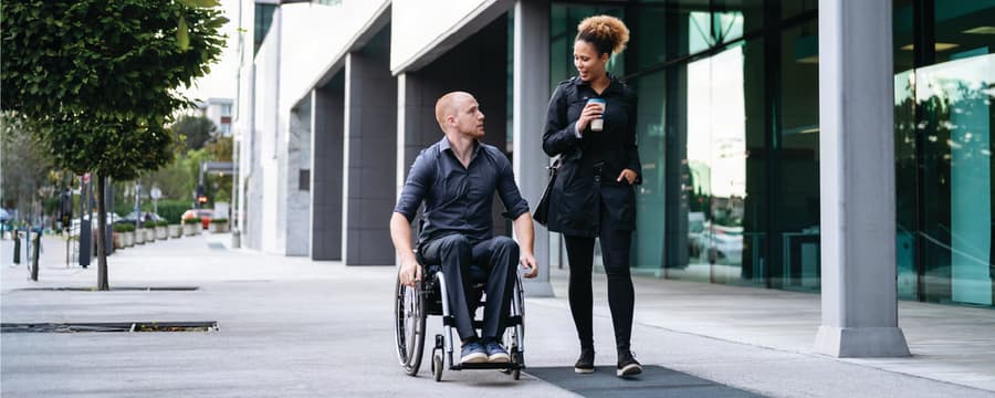 Man using a wheelchair talks with a woman holding a coffee as they walk together outside a modern office building.