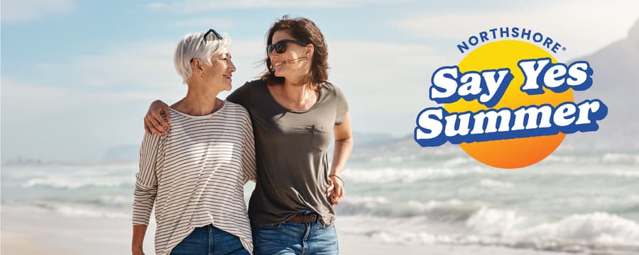 Two women walk arm in arm along a sandy beach, smiling at each other as waves roll in behind them on a sunny day. The upper-right corner features the NorthShore "Say Yes Summer" campaign logo in blue and orange.