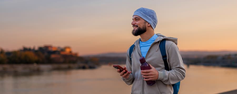A bearded man wearing a blue beanie and gray jacket stands outdoors at sunset, holding a smartphone and water bottle with a backpack on his shoulders.