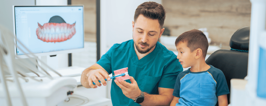 A dental professional in teal scrubs demonstrates proper toothbrushing technique using a large model of teeth and a toothbrush while a young boy watches attentively in a dental exam room. A monitor displaying a dental image is visible in the background.