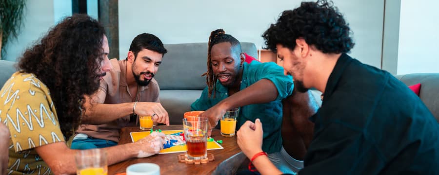 Four friends gathered around a wooden table, smiling and playing a colorful board game while enjoying drinks.