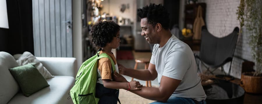 Father smiling and helping his young child put on a bright green backpack before heading out.