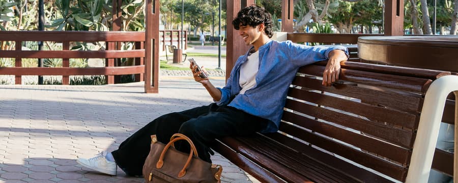 Young man sitting on a park bench, smiling while looking at his smartphone with a travel bag beside him.