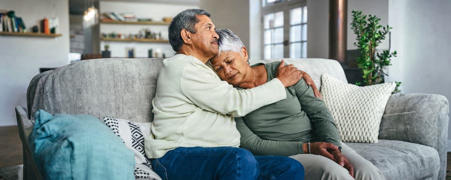 Older man gently embraces and comforts an older woman resting her head on his shoulder while sitting together on a living room couch.