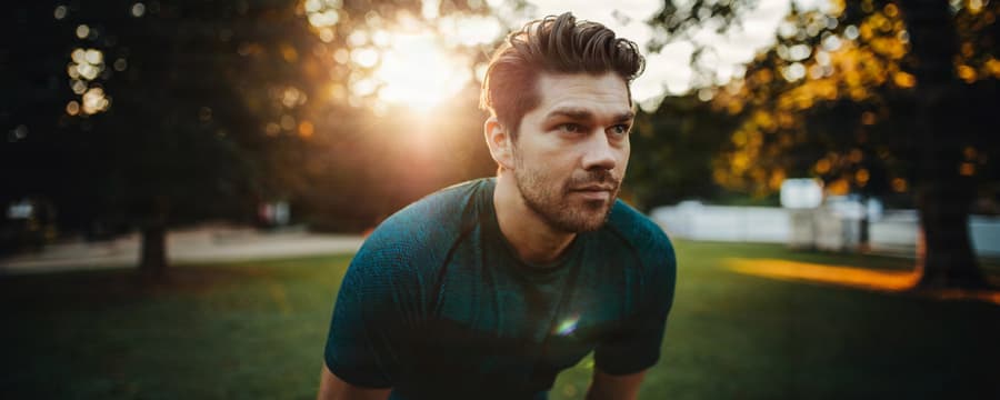 Athletic man wearing a dark teal shirt leaning forward outdoors in a grassy park, with warm sunlight filtering through trees behind him.