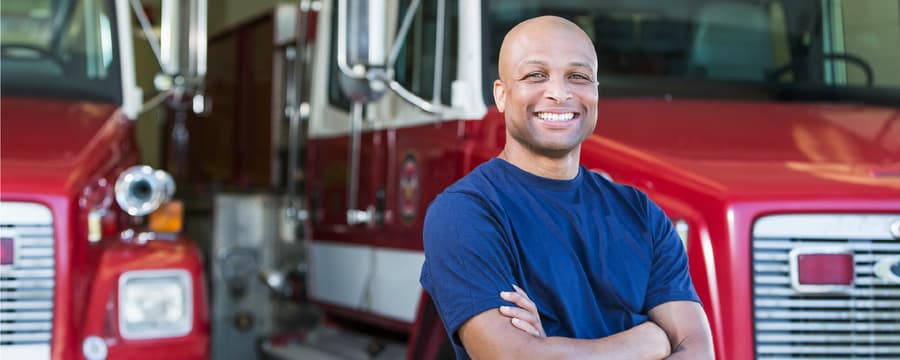 Smiling man in a navy blue T-shirt stands confidently with arms crossed in front of two red fire trucks at a fire station.