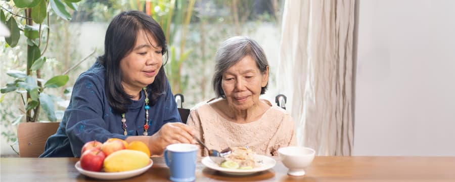 A woman assists an elderly woman at a table, helping her eat a meal in a bright room with greenery visible outside.