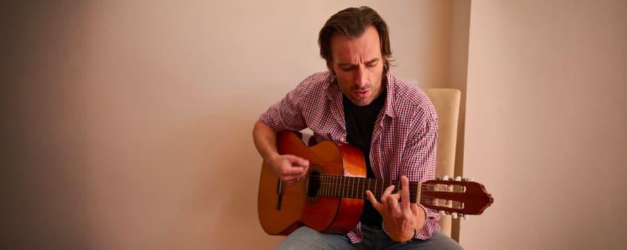 Man seated indoors playing an acoustic guitar, wearing a red plaid shirt over a black T-shirt.
