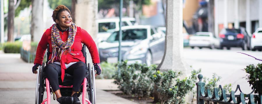A smiling person using a wheelchair moves along a city sidewalk lined with trees and low shrubs. They are wearing a bright red coat, a colorful patterned scarf, dark pants, and gloves. Parked cars and urban buildings appear blurred in the background.