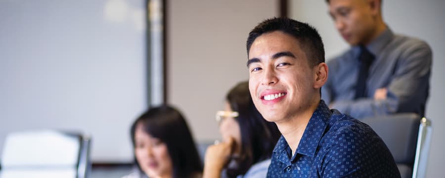 A young man in a blue patterned shirt smiles at the camera while sitting at a desk in a meeting room. Other people in the background are working on laptops and talking, suggesting a collaborative office or classroom setting.