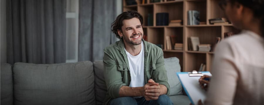 A smiling man in a green shirt sits on a couch during a healthcare consultation with a provider in a white coat, who holds a clipboard. Warm wooden shelving lines the background of the clinical office setting.
