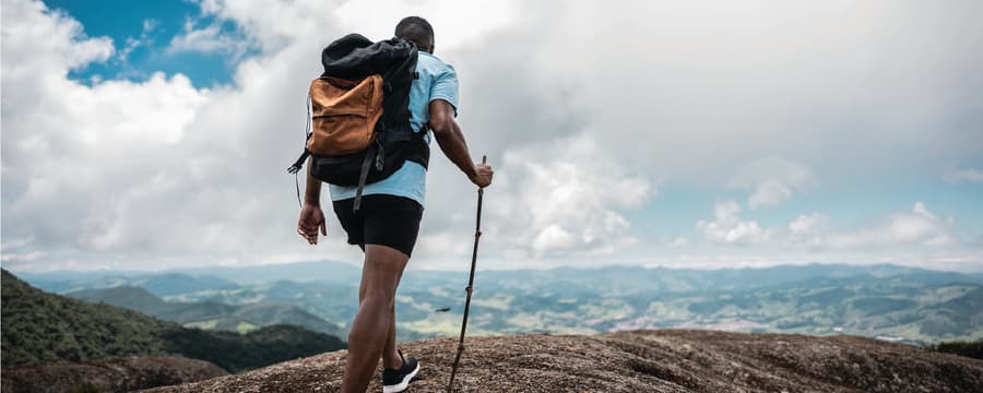A hiker with a large backpack stands on a rocky mountaintop, holding a walking stick and looking out over a wide landscape of hills under a partly cloudy sky.