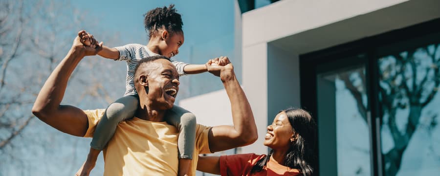 A joyful family outdoors on a sunny day, with a man carrying a young child on his shoulders while smiling, as a woman stands beside them laughing and reaching toward the child.