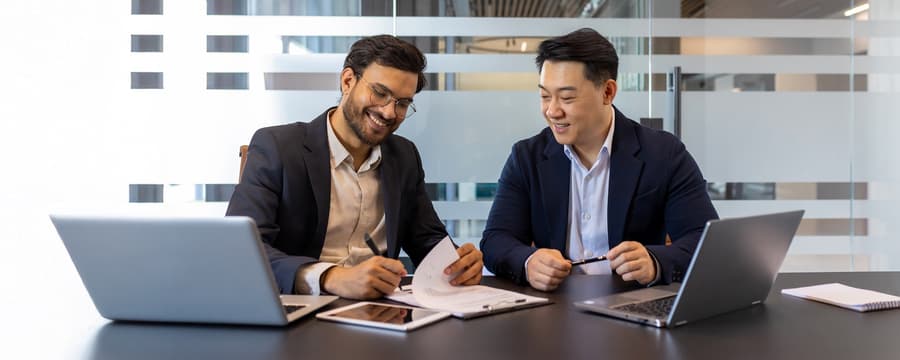 Two businessmen in a modern office setting, reviewing documents and working together at a table with laptops and a tablet.