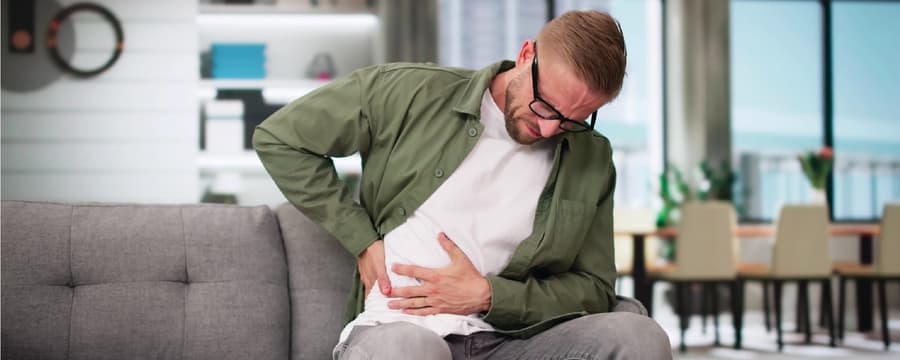 A man sitting on a couch grimaces in pain while clutching the lower right side of his abdomen with one hand and supporting his back with the other, suggesting abdominal or side pain in a home living room setting.