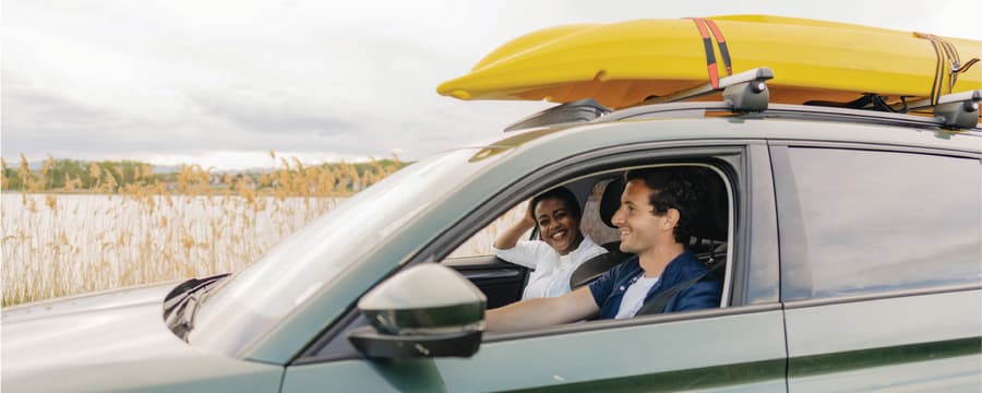 Two people smile from the front seats of a green SUV with two yellow kayaks secured to the roof rack, parked beside a lakeshore with tall reeds in the background.