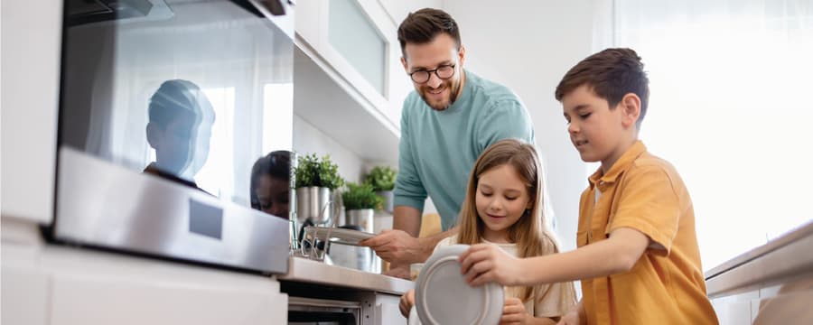 A man and two children load dishes into an open dishwasher in a bright, modern kitchen, smiling and working together.