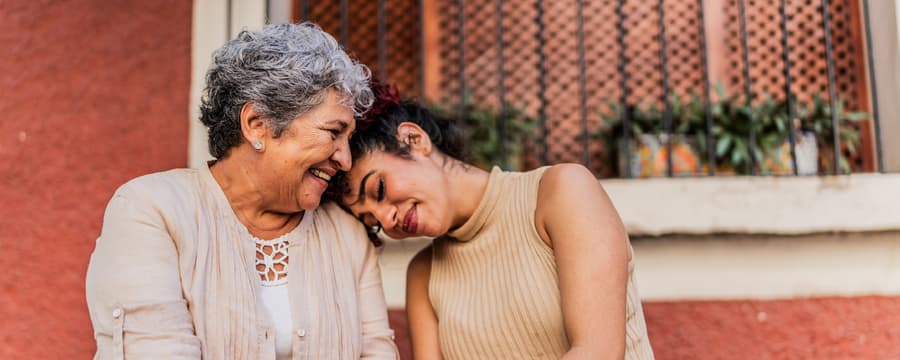 Two women, an older woman and younger woman, sitting together against a terracotta-colored wall. They are holding hands, leaning close with their heads together in an intimate, caring moment. Both are smiling warmly. Soft green foliage is visible in the background.