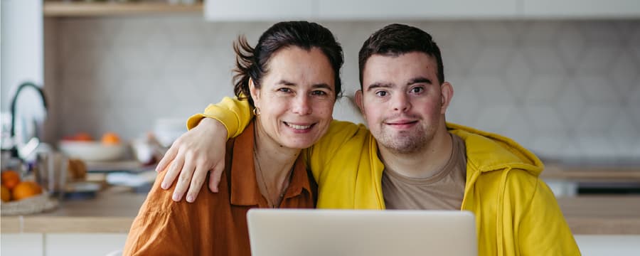 Woman and young man sitting close together at a kitchen table, smiling at the camera behind an open laptop.