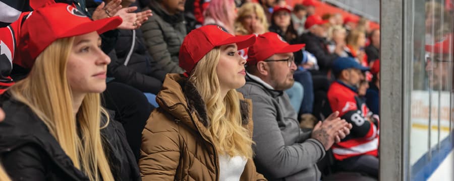 Spectators wearing red caps watch and applaud from the stands at a sporting event.