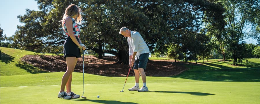 Two people stand on a golf green preparing to putt on a sunny day, with large trees and rolling grass in the background.