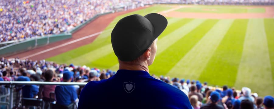 A man wearing a baseball cap sits in the stands, watching a game on a crowded baseball field.
