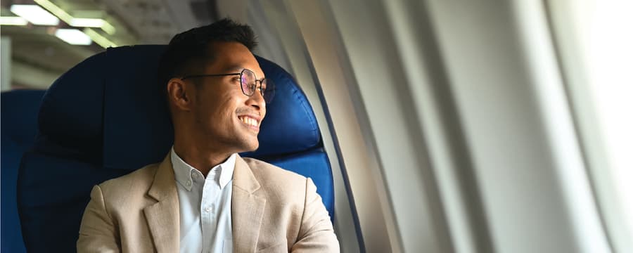 A smiling man wearing glasses and a beige blazer sits by an airplane window, looking outside while resting his hands together on the armrest.